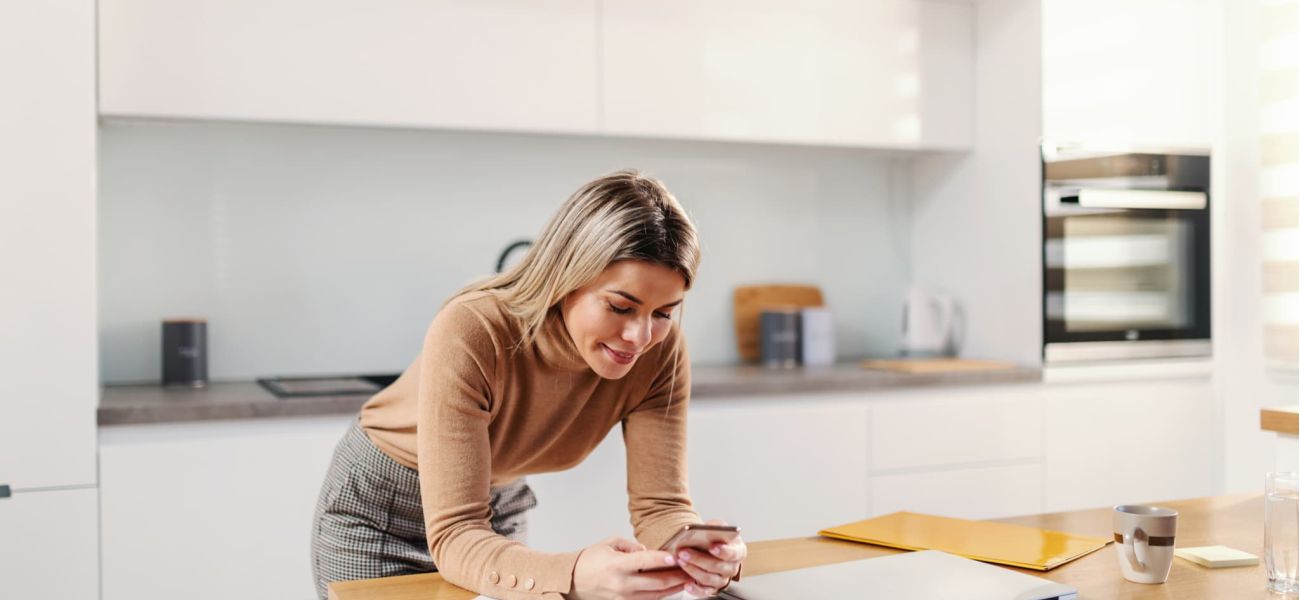 mujer rubia atractiva joven que inclina mesa cocina que usa teléfono inteligente