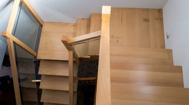 Vista de una escalera de madera en haya con forma en U, con peldaños de madera maciza en tono natural y barandilla de cristal templado.